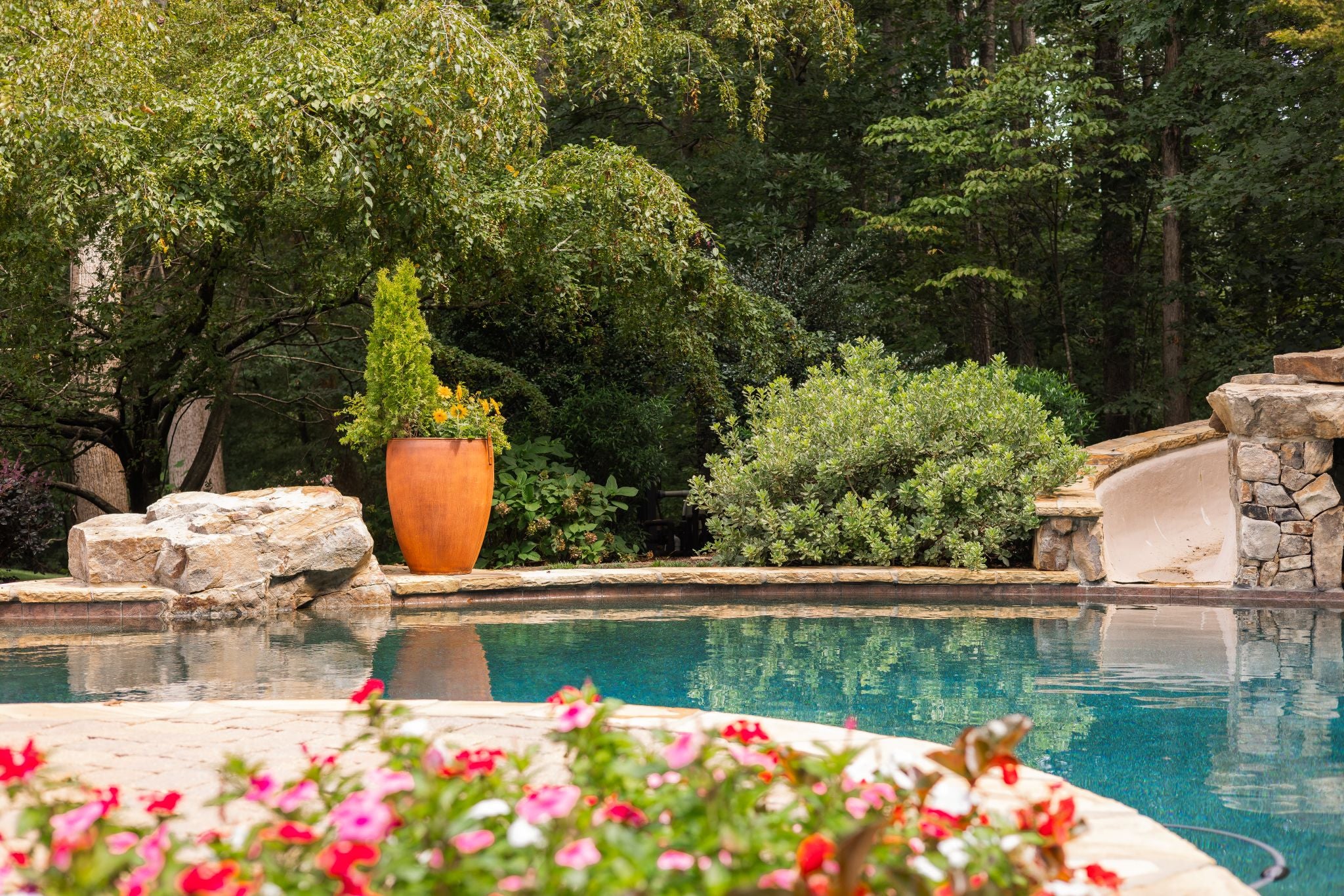 Outdoor pool area with flowers, a large outdoor metal pot, and trees in the background