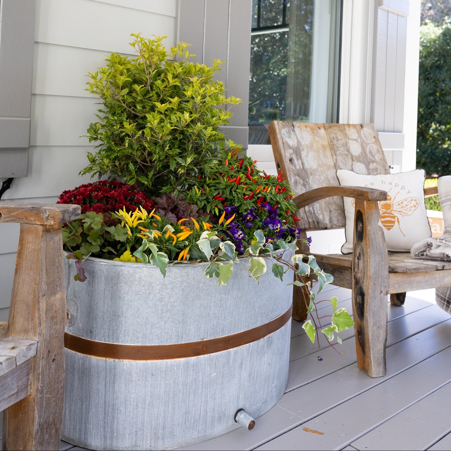 Decorative planter with flowers on a porch with a wooden chair and cushion.