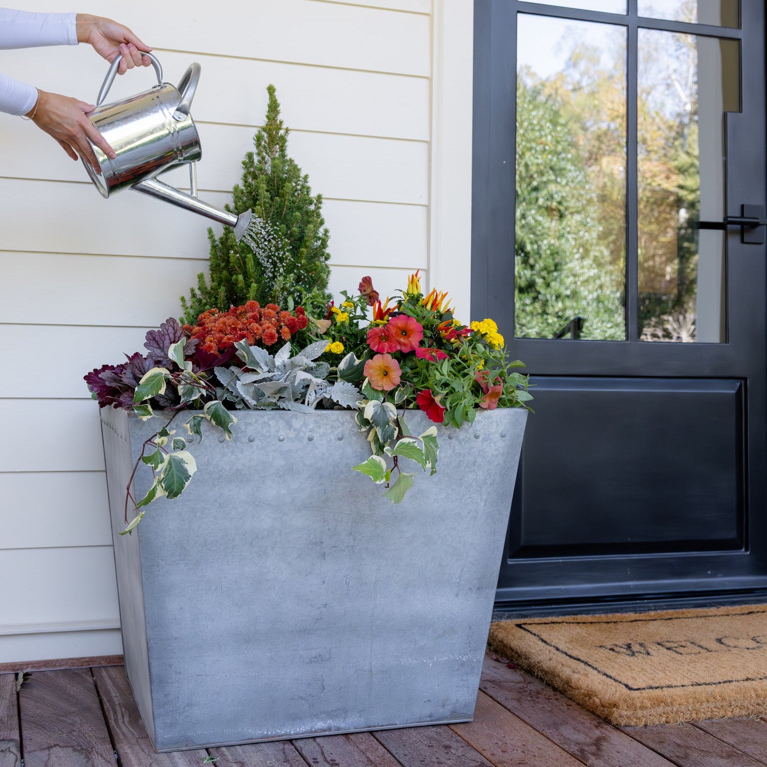 Person watering a large metal planter with flowers on a porch.