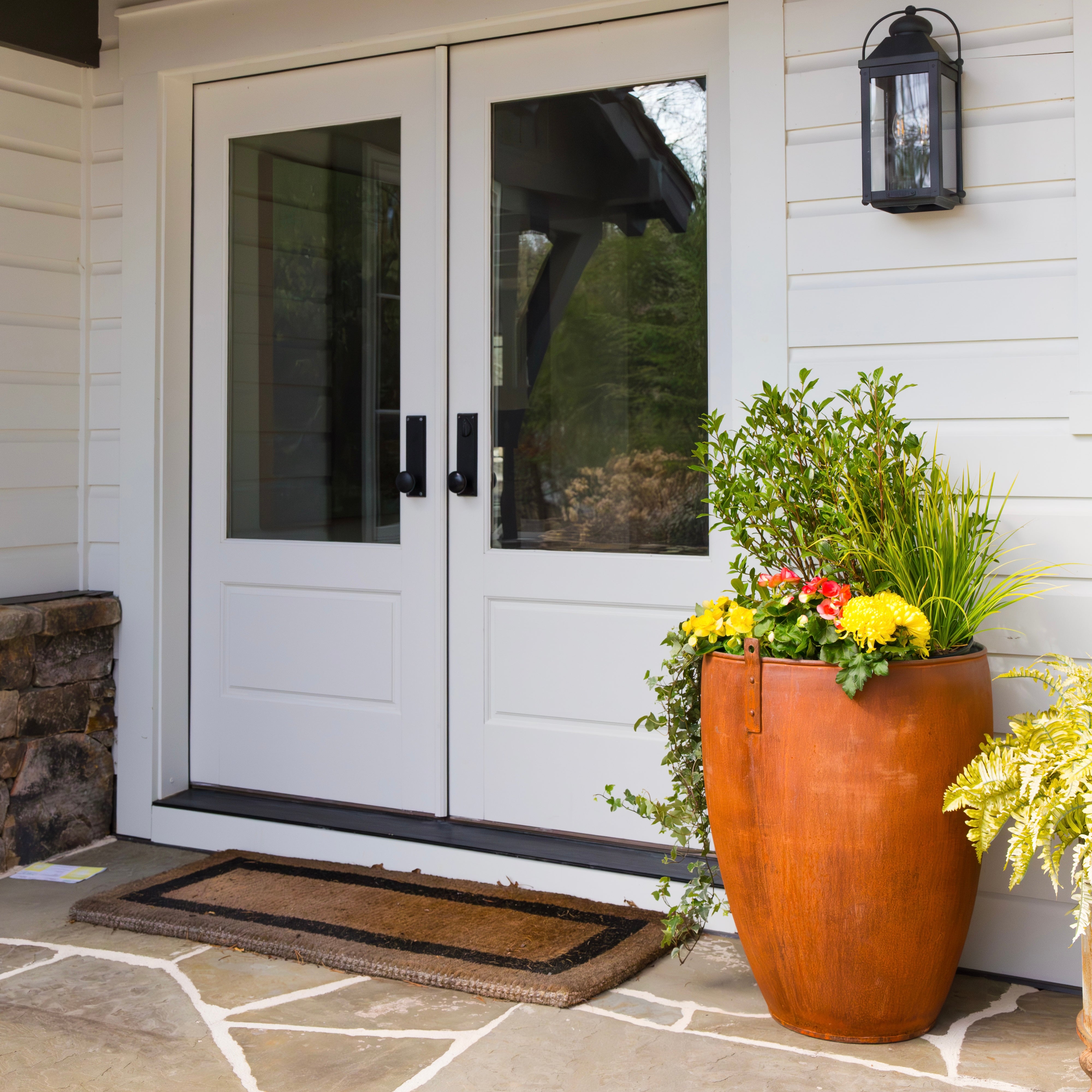Front door of a house with a doormat, potted plants in large steel planter, and a lantern.