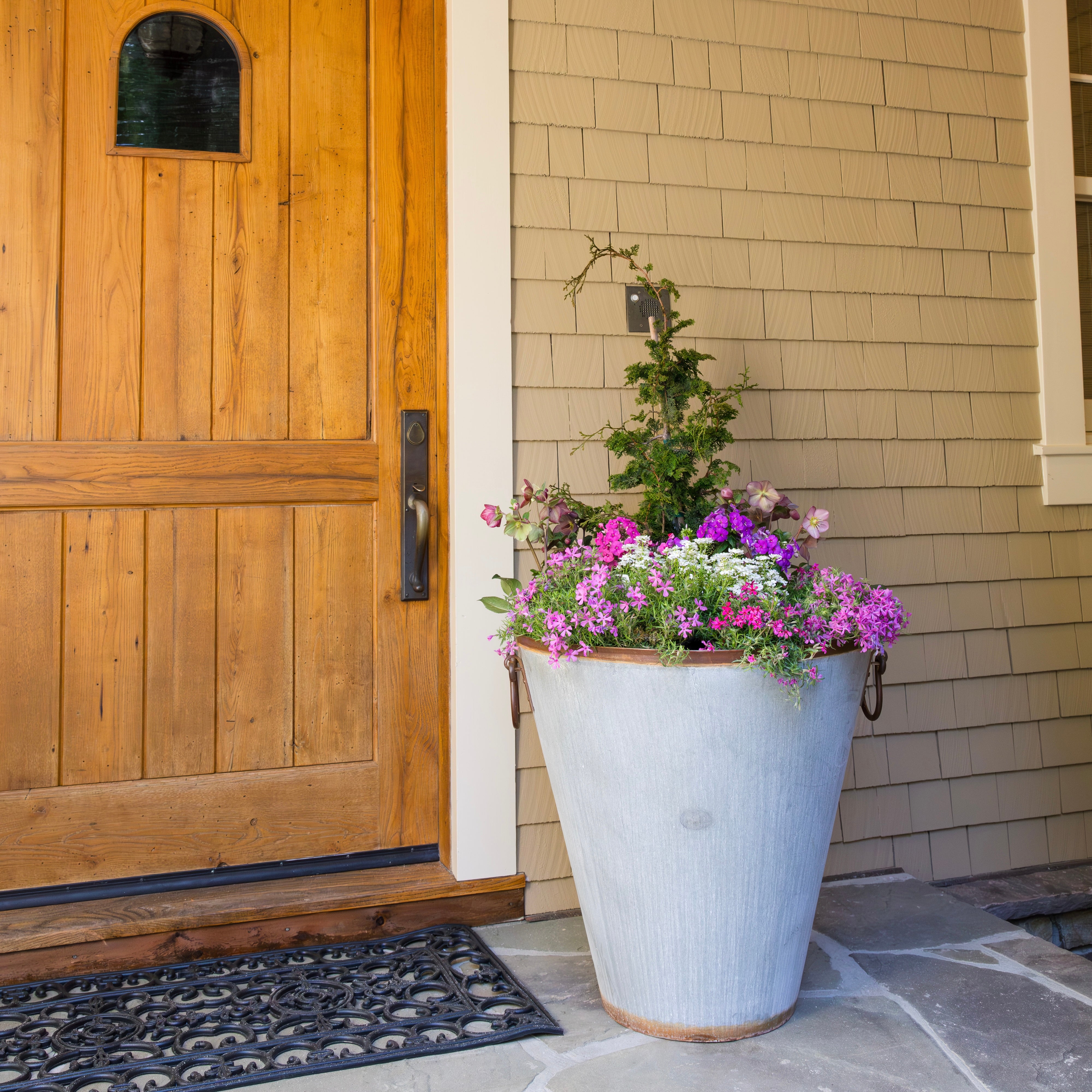 Floral arrangement in a large steel pot on a porch with a wooden door and beige wall.