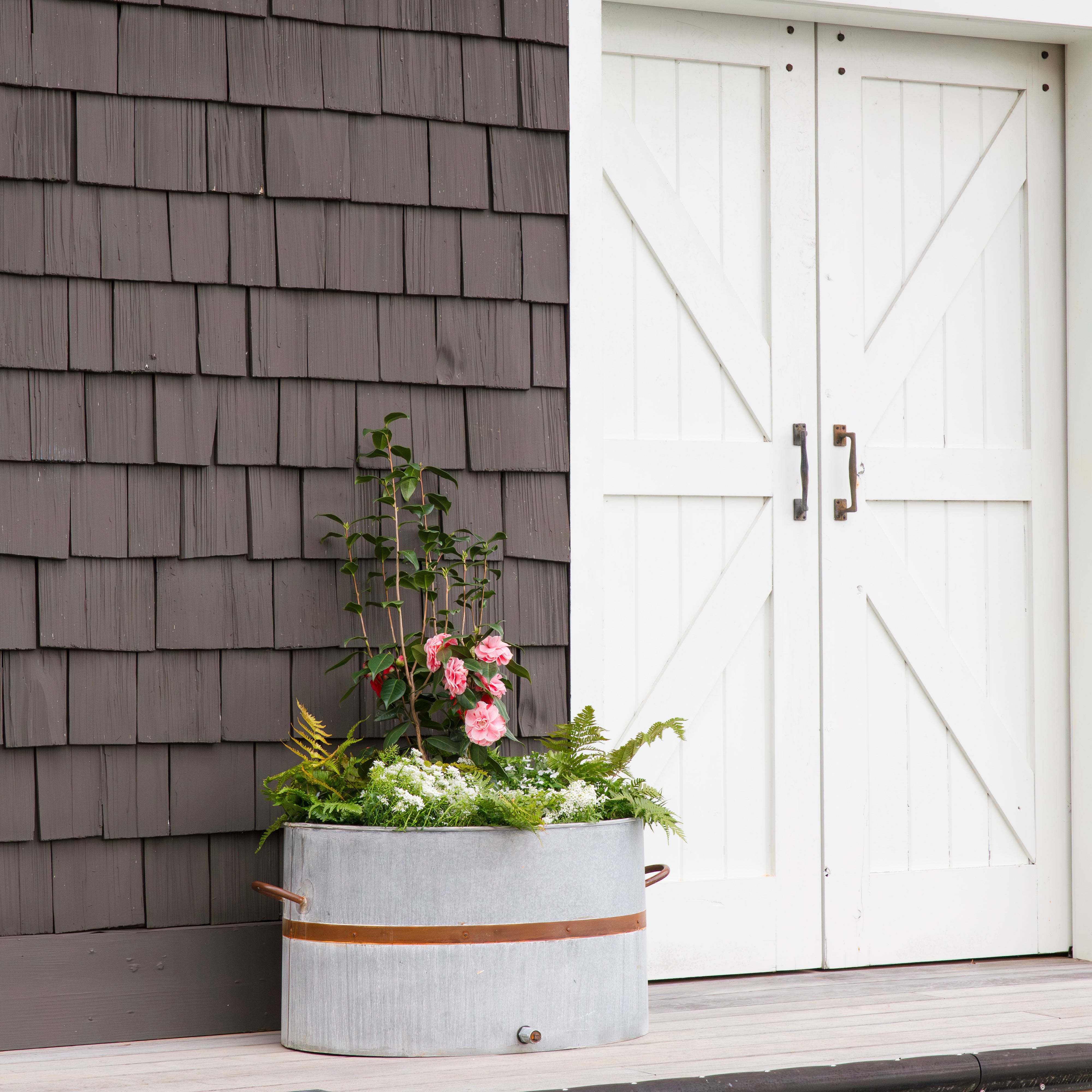 Decorative planter with flowers in front of a gray shingled wall and white doors.