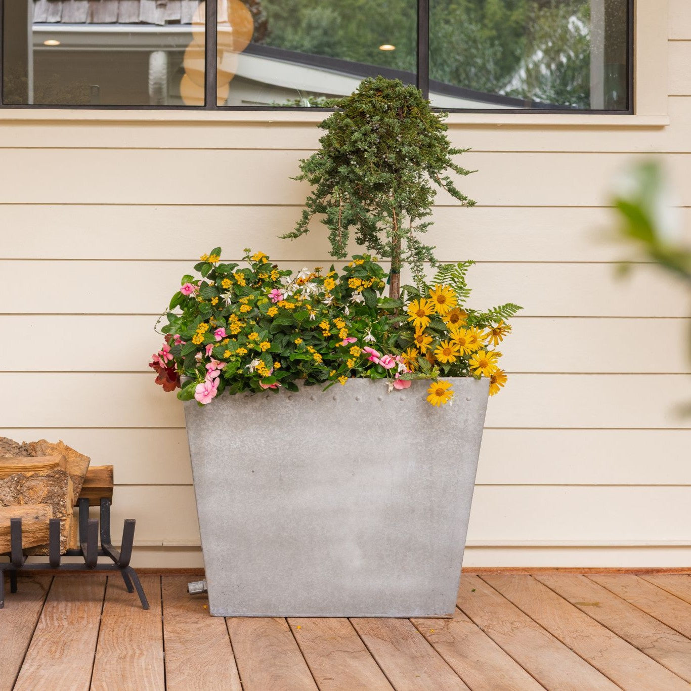 Decorative large planter with flowers and a tree on a wooden deck against a beige wall.