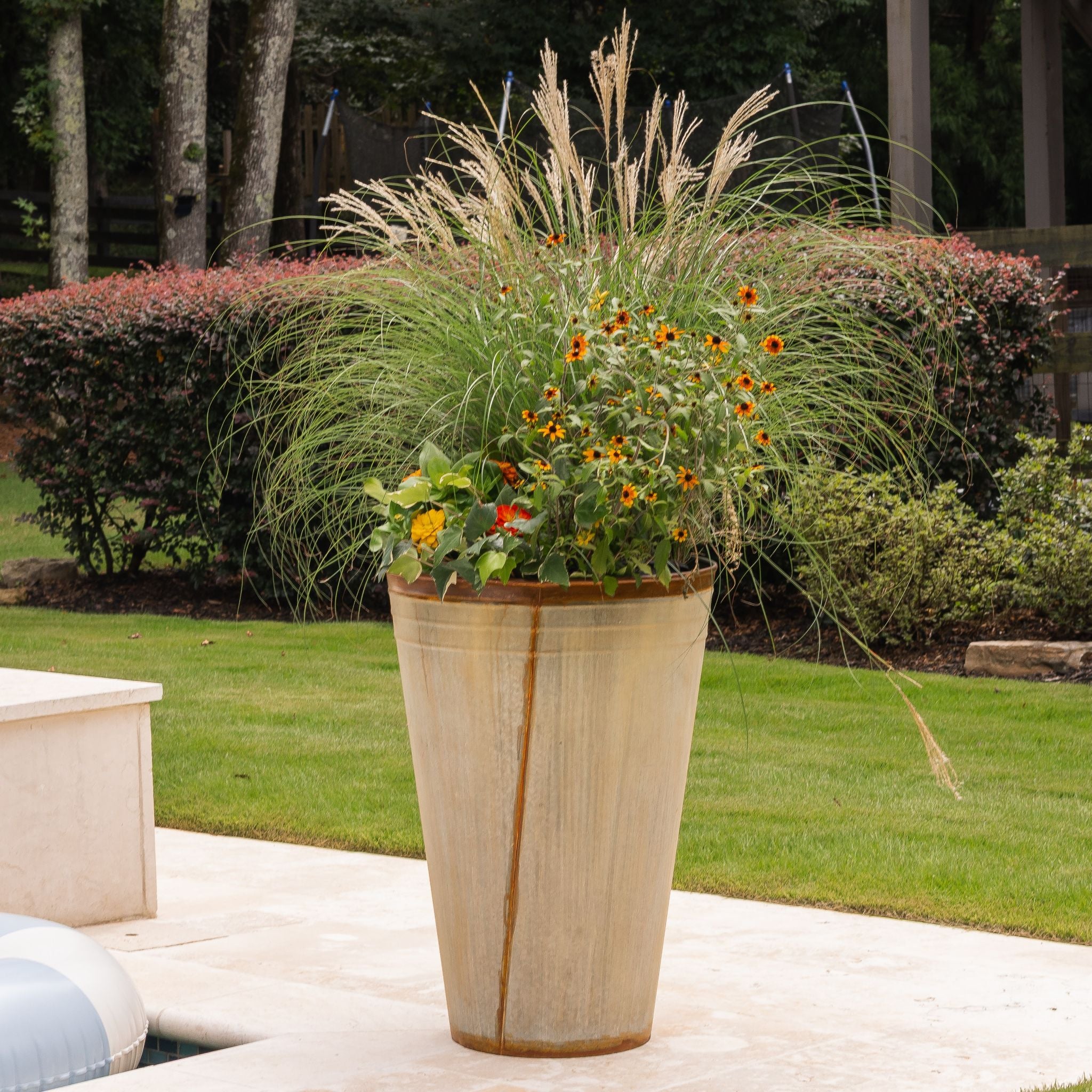 Tall decorative planter with grasses and flowers on a pool patio.