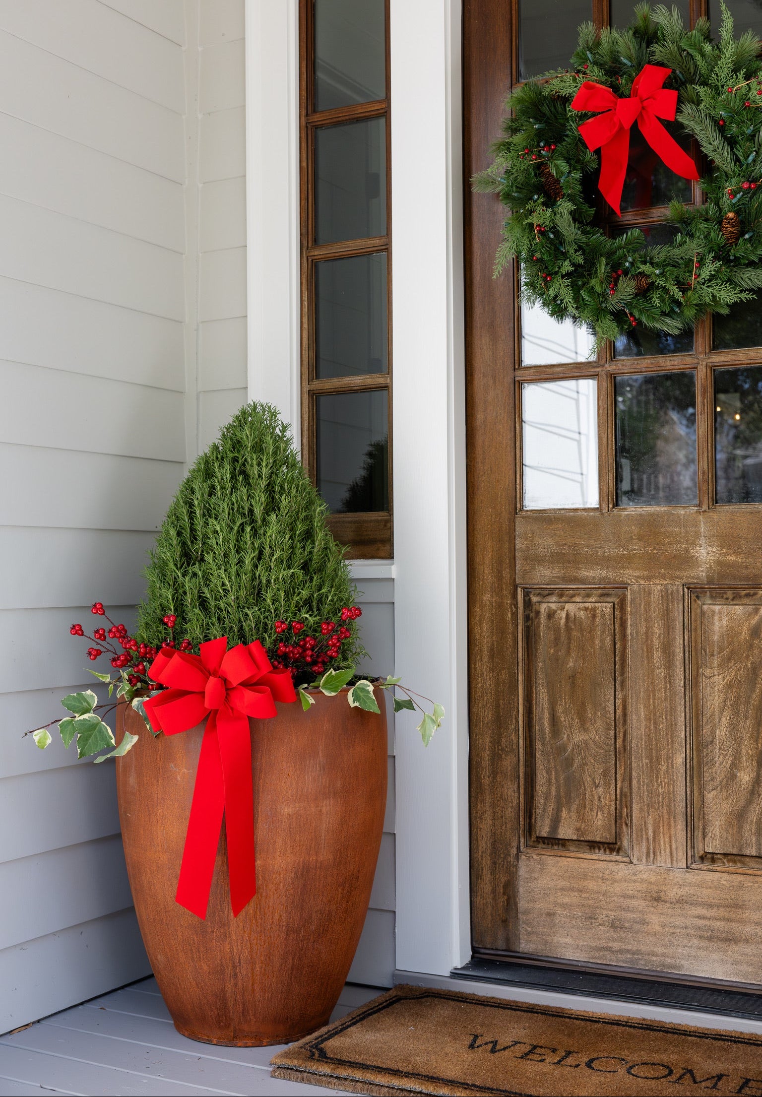 Decorative front door area with a Christmas wreath and a large potted metal planter with red bows. 